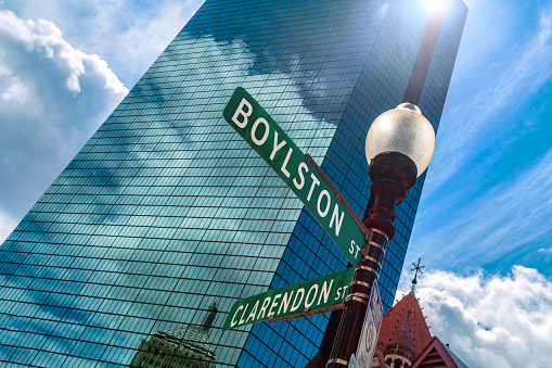 Boston, Massachusetts, USA - July 30, 2023: Close-up of Boylston and Clarendon street signs on a street light pole in Copley Square, a public square in Boston's Back Bay neighborhood. In the background is the 200 Clarendon Street skyscraper, formerly named the John Hancock Tower, and a small portion of Trinity Church. The reflection of clouds and the old John Hancock building is in the blue glass of the Hancock Tower. The Hancock Tower is highest building in Boston and all of New England. Lens flare.