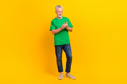 Full length portrait of cool cheerful aged person hold use smart phone typing blog post isolated on yellow color background.