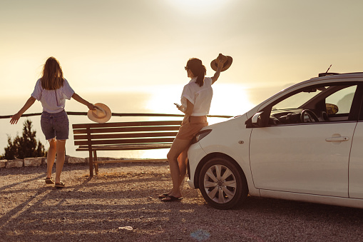 Rear view of two young women taking a break during a road trip. They are standing on a viewpoint and looking at the sunset
