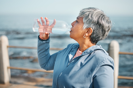 Outdoor, fitness or senior woman drinking water for hydration, wellness and thirsty on beach run, training or exercise. Bottle, runner or elderly person refresh with cold liquid on retirement workout