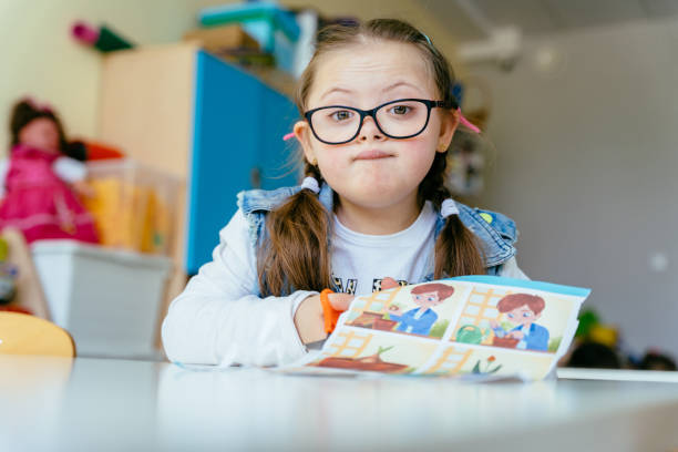 istock Cute little junior schoolchild with special needs having funny emotion on her face sits at desk at classroom works independently. 1598626443