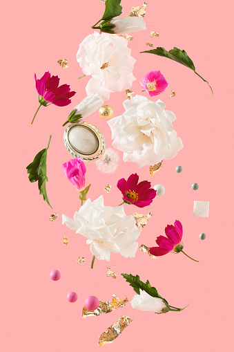 Creative composition of levitating white roses, hibiscus buds, rose evening primroses, magenta cosmos flowers, golden flakes, white, pink, golden, turquoise beads, green leaves, dandelion and baroque brooch over pastel pink background.