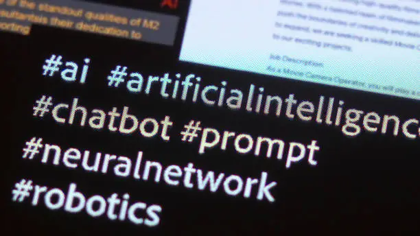 Stock image; an extreme close-up of a smart phone screen showing hashtags on a dark screen. They’ve spelling out AI; artificial intelligence; chatbot; prompt; neural network and robotics. Ie:  #ai  #artificialintelligence  #chatbot  #prompt #neuralnetwork  #robotics.