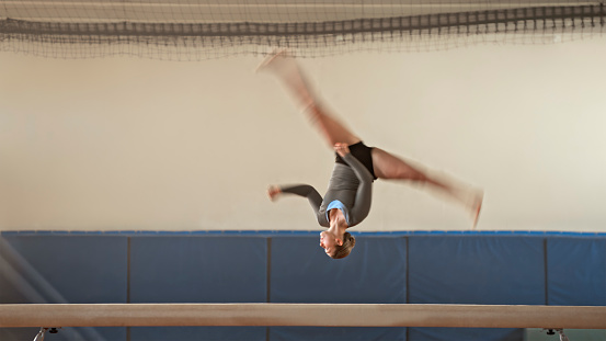 Female gymnast performing backflipping on balance beam during practice in sports hall.