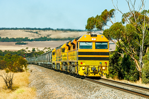 Rail infrastructure serving the farming and food production industries: four powerful diesel locomotives work a loaded grain train through rolling hilly landscape on way to port. In the distance are a range of hills and a red railway signal.  ID and Logos edited.