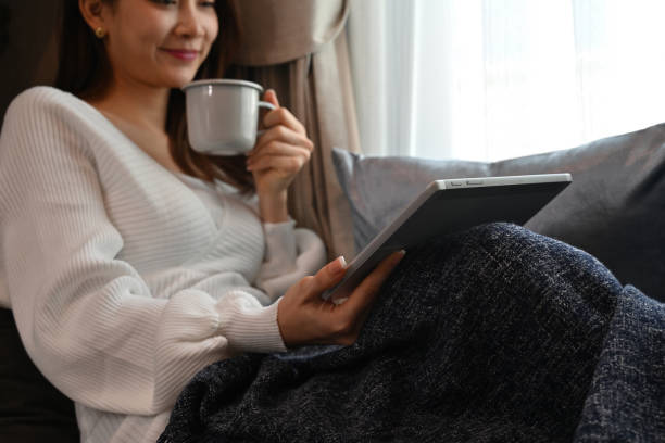 Cropped image an Asian woman sitting with legs stretched and covered with a blanket while working on a digital tablet and enjoy with hot drink next to the window.