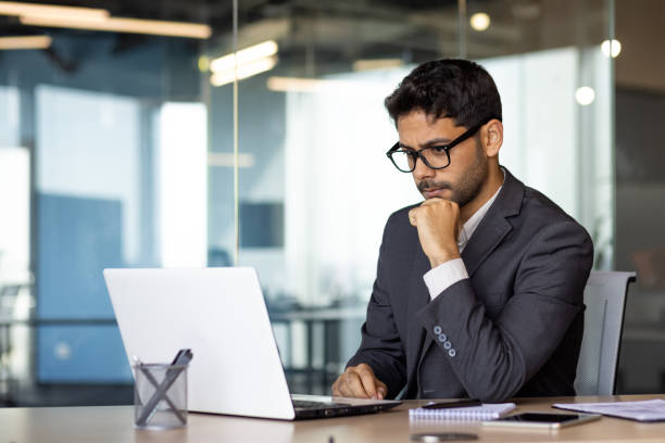 istock Young thinking businessman at workplace with laptop inside office, boss in glasses and business suit thinking about technical financial solutions, resting hand on chin 1598829404