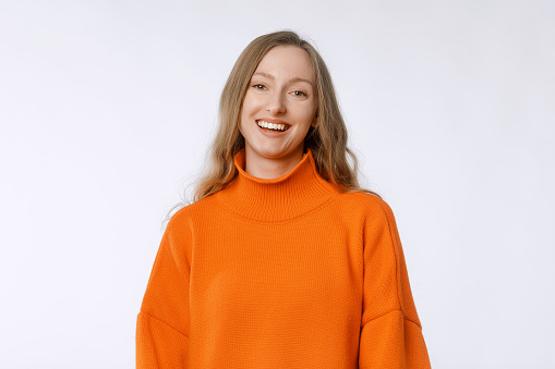 Portrait of smiling cheerful girl, looks happy and upbeat at camera, stands in trendy knitwear orange sweater against neutral studio background