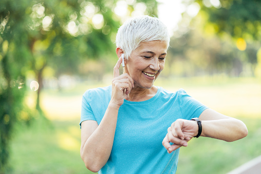 Senior woman tired after jogging with sport clothing and sport technologies. Mature female athlete relaxing with great music in headphones before sport training in the park