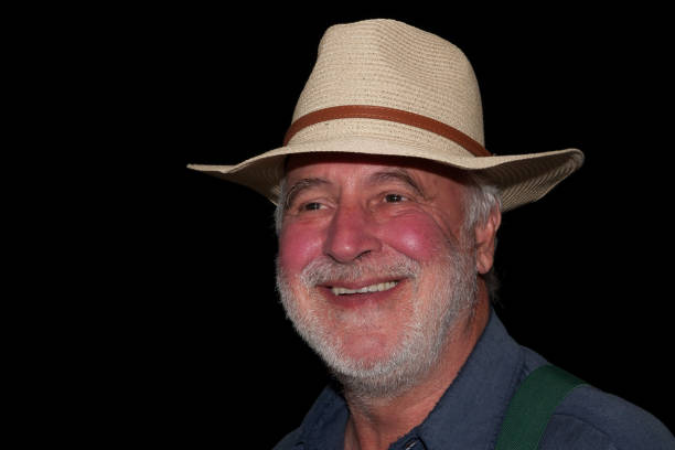 Portrait of a funny senior farmer with a straw hat laughing heartily at the camera against a black background.