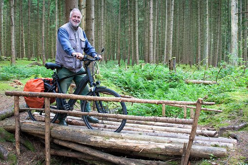 Deep in the forest, away from the bike paths, a senior pushes his bike over a bumpy forest bridge made of logs.