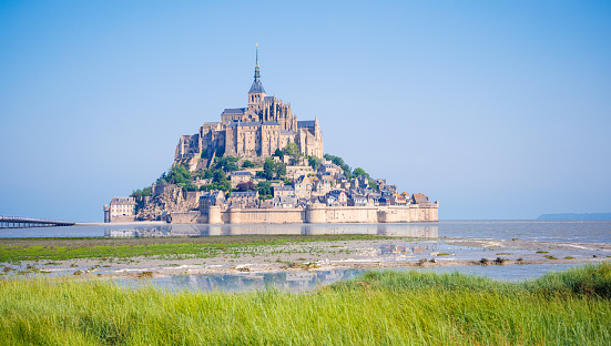 Normandy, France, August 29, 2019: Looking at the magestic Mont Saint-Michel  island during low tide, it makes the city look like it was built on dry sand.  Le Mont-Saint-Michel or in english Saint Michael's Mount is a tidal island and mainland commune in Normandy, France. The island lies approximately one kilometre (0.6 miles) off the country's north-western coast, at the mouth of the Couesnon River near Avranches and is 7 hectares (17 acres) in area. The mainland part of the commune is 393 hectares (971 acres) in area so that the total surface of the commune is 400 hectares (988 acres). As of 2017, the island has a population of 30. \nAs of 2017, the island has a population of 30. Mont-Saint-Michel and its bay are on the UNESCO list of World Heritage Sites. It is visited by more than 3 million people each year. Over 60 buildings within the commune are protected in France as monuments historiques.