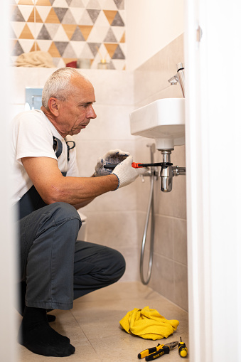 In this professional scene, a mature plumber wearing a uniform is shown diligently fixing a sink using various work tools. With focused attention and expertise, the plumber works to resolve any issues and restore proper functionality to the sink.