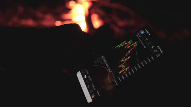 Businessman Using Mobile Phone For Checking Stock Chart At Night With Camping Bonfire In Background