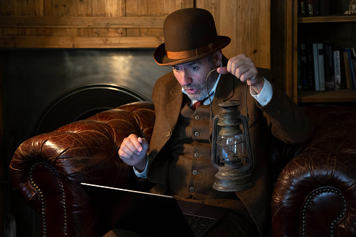 A man in a hat and a Victorian-era costume with an antique lamp in his hand looks with surprise at the laptop in a retro style. The theme is modern technology and the last century.