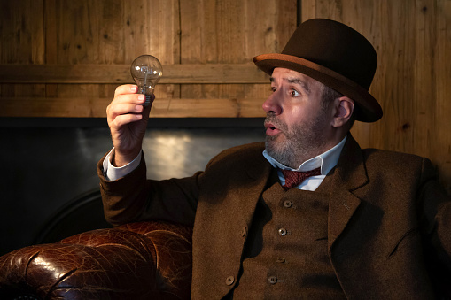 A man in a Victorian hat and suit is looking at a light bulb in his hand in a retro-style salon. The theme is electricity in the last century and science in history.