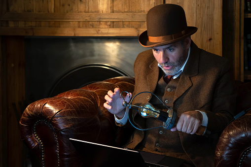 A man in a hat and a Victorian-era costume with an antique lamp in his hand looks with surprise at the laptop in a retro style. The theme is modern technology and the last century.