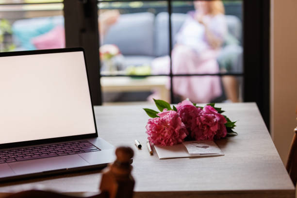 Selective focus shot of a laptop with white, blank screen, bouquet of flowers and a greeting card next to it.