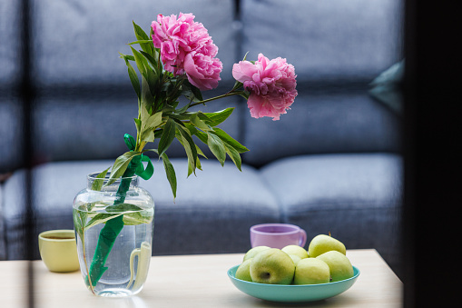Selective focus shot of a bouquet of pink peonies in a vase as decoration next to a bowl of green apples served on a coffee table.