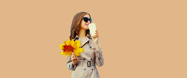 Beautiful young woman with yellow maple leaves and cup of coffee looking away wearing coat, sunglasses on brown wall studio background