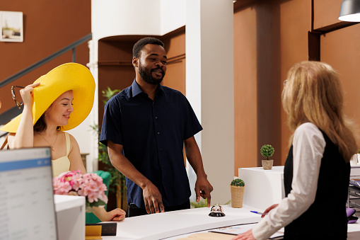 Multiethnic couple smiling and talking with friendly receptionist at front desk of exotic beach resort about their honeymoon trip. Hotel employee helps clients to register at reception counter.