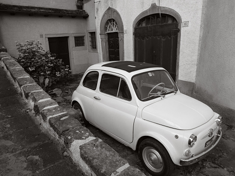 Fiumalbo, Italy - July 25, 2023: A white Fiat 500 is parked on a cobblestone street. The car is in front of a pink building with white trim, which has a wooden door and two windows with white shutters. On the left side of the street, there is a small garden with green bushes and a stone wall.