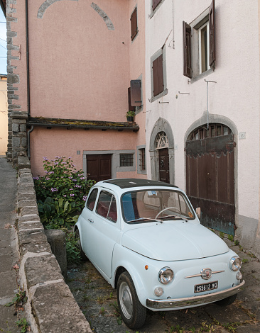 Fiumalbo, Italy - July 25, 2023: A light blue Fiat 500 is parked on a cobblestone street. The car is in front of a pink building with white trim, which has a wooden door and two windows with white shutters. On the left side of the street, there is a small garden with green bushes and a stone wall.