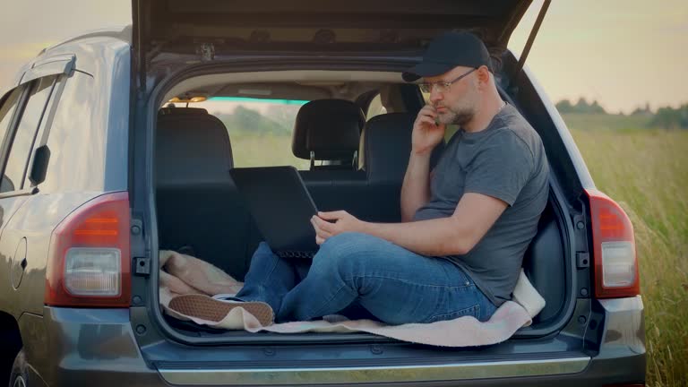 Technician engineer Man working remotely on laptop looking thoughtfully thinking about hard project in nature sitting in car trunk while traveling