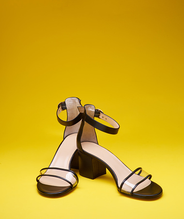 A pair of feminine open-toe shoes with block heels, clear vamps, ankle straps, and beige insoles, isolated on a bright yellow background. Concept for a modern and fashionable shoe store.