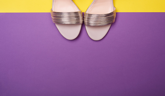 Shiny summer golden metallic women's sandals on a purple-yellow background with large copy space on bottom. Flat lay, top view. Fashion blog, summer season footwear concept