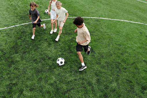 Above angle of several schoolchildren playing soccer on green football field at stadium while three of them running after ball during game