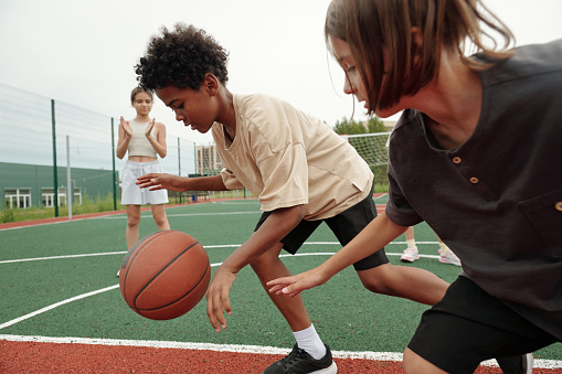 African American schoolboy in activewear dribbling ball while running forwards in front of his classmate while blond girl standing on background
