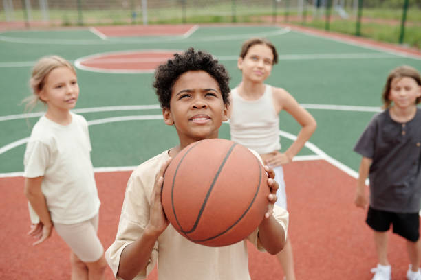 istock Cute schoolboy with ball looking upwards before throwing it into basket 1599099882