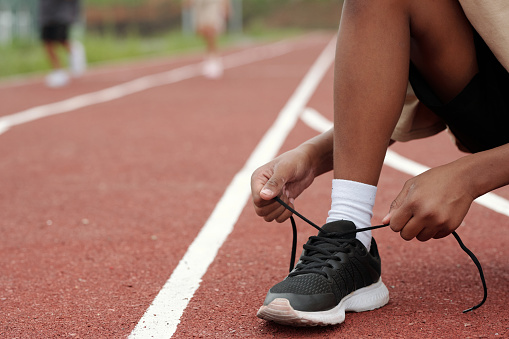 Leg bent in knee of African American schoolboy tying shoelace of sneaker while sitting on squats on race track at stadium during training
