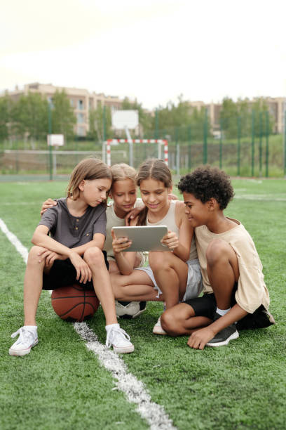 Group of active boys and girls sitting on football field and using tablet Group of active boys and girls sitting on football field and using tablet while watching online video or searching for one in the internet 9 stock pictures, royalty-free photos & images