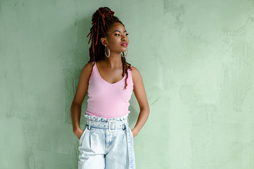 Portrait of gorgeous African girl with dreadlocks hairstyle wearing wide-leg jeans and pink top, looking away, isolated on light green wall with copy space. Beautiful confident female model.