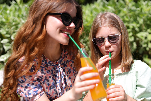 Two cute children drinking orange soda from glass bottles in a park. They wear colorful shirts and enjoy their summer day with green straws.