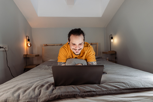 Portrait of a smiling Cuban man using a laptop while lying on the bed. Evening at home.