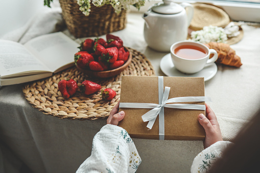 The child's hands hold a beautiful gift box with a ribbon against the backdrop of a festive breakfast. Top view, close-up. Happy mother's day.