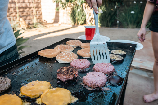 Multiple burger patties, buns, and zucchinis being grilled at a summer barbecue party in LA in the day.