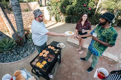 A man wearing a white t-shirt and bucket hat hands a plate with a cheeseburger to his friend in a green tie dye shirt.