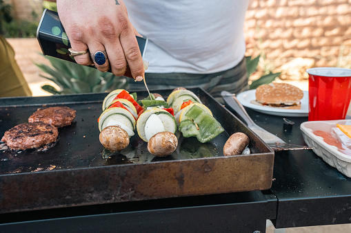 Close-up shot of a man dousing cooking oil on a skewer with vegetables; bell peppers, zucchini, mushroom, onion, and tomatoes, while cooking burgers atop a grill.
