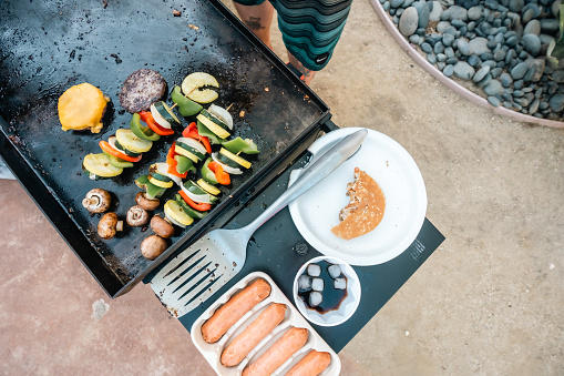 An overhead shot of a grill with vegetable skewers and burger patties. Next to the grill, a half-eaten hamburger, a cup of soda, and packed hotdogs.