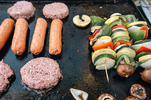 A close-up shot of four burger patties, four sausages, and three vegetable skewers cooking on a grill.