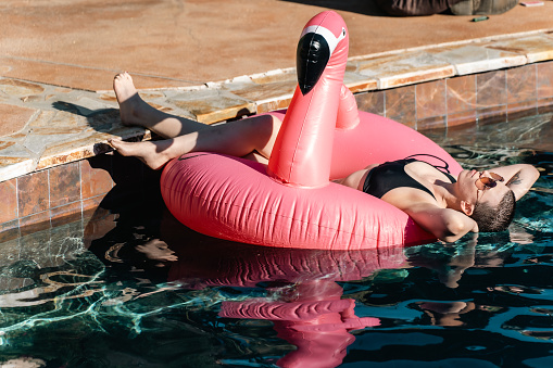 A non-binary person in black swimming attire laying on a pink inflatable float shaped like a flamingo; lazing around by the poolside.