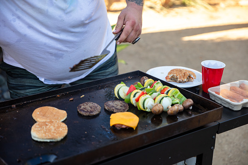 A man wearing a white t-shirt holding a spatula in front of a grill with vegetable skewers and burger patties. Next to the grill, a half-eaten hamburger, a cup of soda, and packed hotdogs.