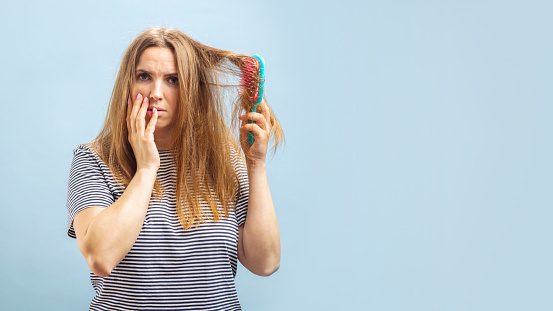 Very sad and upset young woman looking with shock at her damaged hair on blue background.