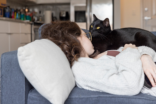 Young brunette curly haired woman in eyeglasses takes a nap with her black cat on the sofa in a bright loft apartment. Woman turned away from the screen