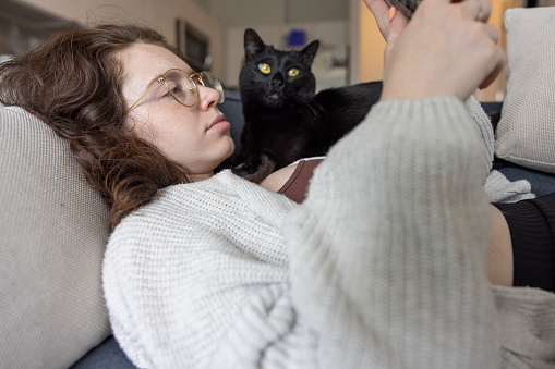 Young woman with glasses uses her smartphone to surf the net as she relaxes with her black cat on the sofa in a well-lit loft apartment