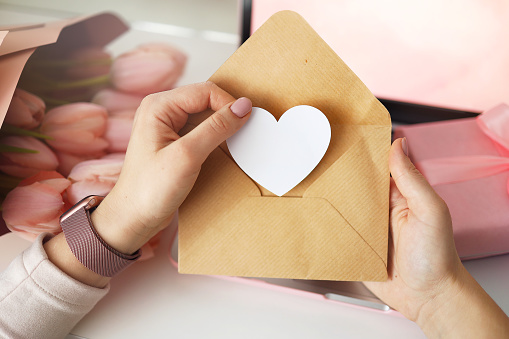 Woman's hands holding a letter in craft envelope. Pink background, valentine's day concept. Tulips flower and pink gift box in background. Womens home desk.